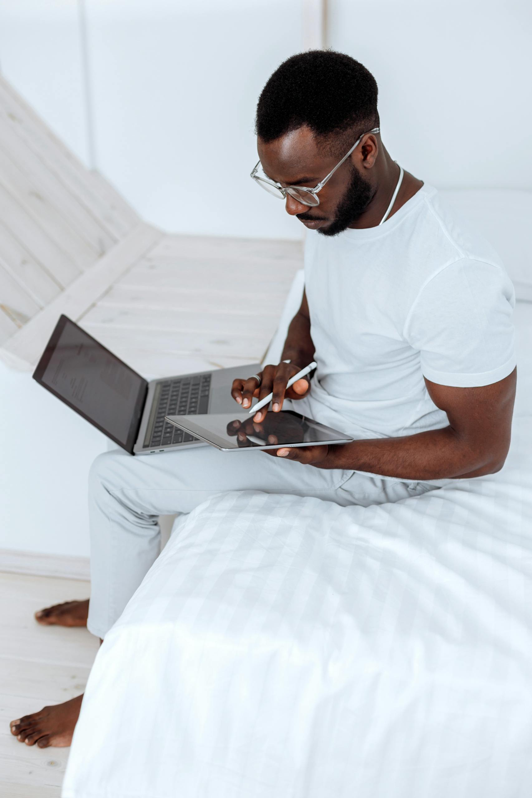 AI Fundamentals Course 1 A man wearing eyeglasses sits on a bed using a laptop and tablet for remote work. He is learning the AI Fundamentals Course at Zindua School