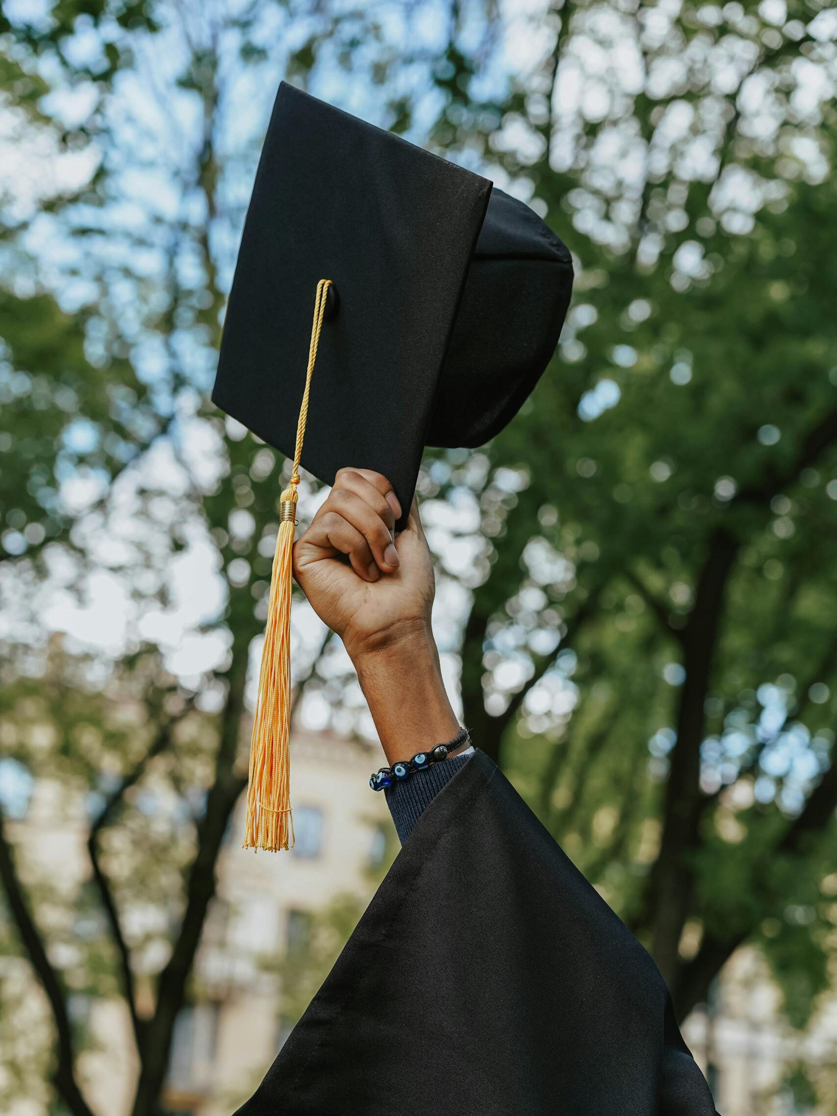 A close-up of a hand raising a graduation cap in celebration among trees.